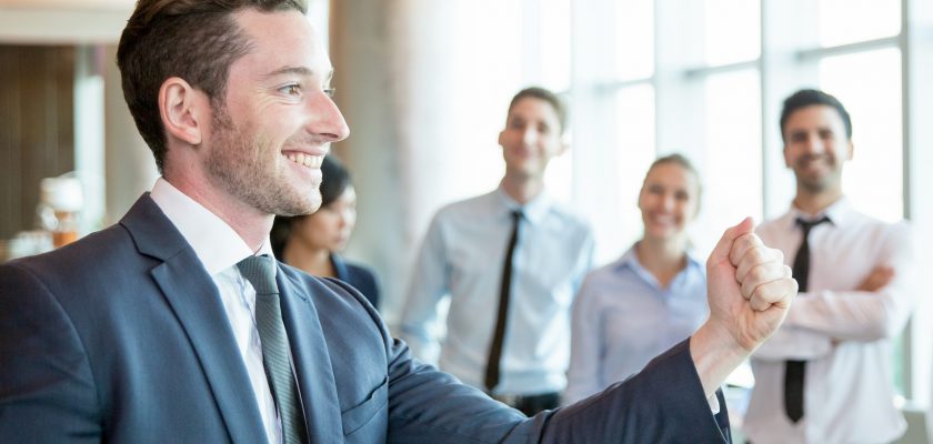 Cheerful leader motivating his business team Cheerful leader motivating his business team. Handsome young politician telling his plan and showing fist as symbol of power. Positive business people standing in background. Strong company concept