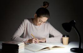 Busy freelancer rewrites infrormation into notepad, prepares article for publication, reads books, writes some notes in organizer, drinks takeaway coffee, sits in darkness, wears round big spectacles