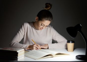 Busy freelancer rewrites infrormation into notepad, prepares article for publication, reads books, writes some notes in organizer, drinks takeaway coffee, sits in darkness, wears round big spectacles