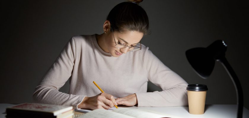 Busy freelancer rewrites infrormation into notepad, prepares article for publication, reads books, writes some notes in organizer, drinks takeaway coffee, sits in darkness, wears round big spectacles