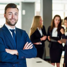 Caucasian businessman leader looking at camera in modern office with multi-ethnic businesspeople working at the background. Teamwork concept. Young man with beard wearing blue suit.