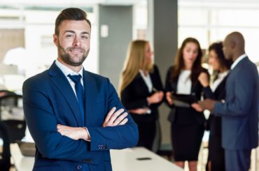 Caucasian businessman leader looking at camera in modern office with multi-ethnic businesspeople working at the background. Teamwork concept. Young man with beard wearing blue suit.